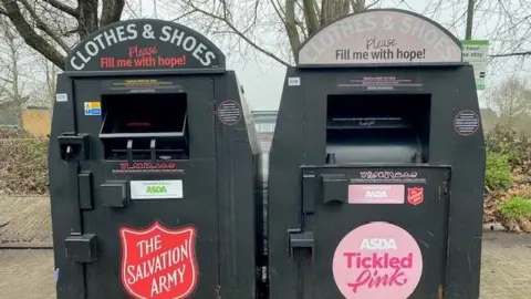 Kate Clayton Two dark green, metal containers with drawers where clothes can be donated. One bears a red logo for The Salvation Army while the other has a pink logo that reads Asda Tickled Pink.