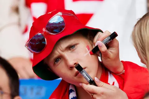 Getty Images A woman paints the Danish flag on a young man's cheek. He is wearing a Danish scarf, a bucket hat and sunglasses
