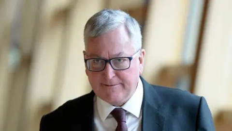 Getty Images Head and shoulders shot of Fergus Ewing. He is wearing a grey suit, white shirt and maroon tie. He has grey hair, combed in a side shed, and is wearing black-rimmed glasses. Ewing has a serious expression on his face.