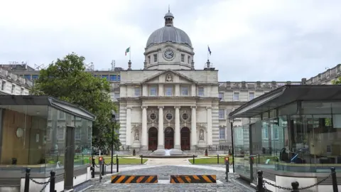 A large white stone building has four large pillars in front of its entrance and a grey dome roof. There is security barriers in front of the waterfountain that sits in front of the building. 