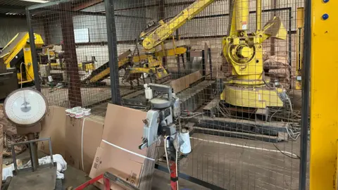 John Devine/BBC Various machinery in a farm barn with unmade cardboard boxes and tape visible, there is also an old fashioned weighing machine on the left.