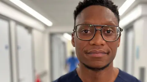 Howie Katyumba, a midwife at Basildon Hospital maternity unit, standing in a corridor, in scrubs and wearing black-framed glasses.