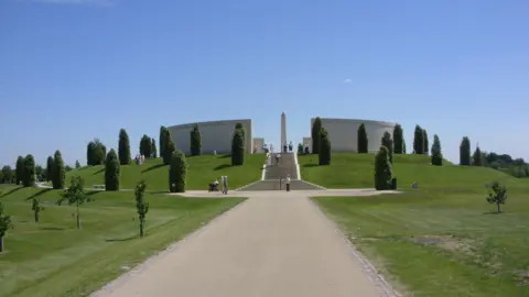 PA Media The main memorial in the centre of the National Memorial Arboretum. It is large, circular and white, with a tall structure at the centre, and is on top of a small hill with steps coming down towards us. It is surrounded by a number of trees.