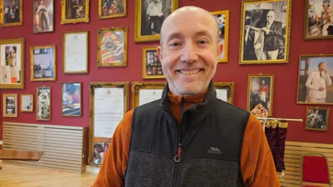 Aisha Iqbal/BBC A man stands smiling. He is aged in his late 50s to 60s and is bald. He wears a burnt orange shirt and brown gilet. He stands in a regal looking room with framed pictures on the walls of royalty, dignitaries and tailors posing with clients and creations.