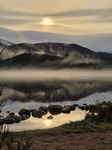 Freya Todd A calm loch reflects a hazy sunrise as mist drifts above the water and low, dark hills rise in the background.