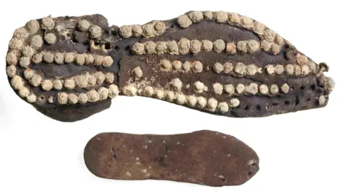 London Museum Roman leather shoes belonging to an adult and a child. The top image shows the adult leather shoe - which looks like the sole of a modern day shoe -  with what looks like several round white studs all over it and the child's shoe is dark brown leather with no similar markings like the adult shoe.