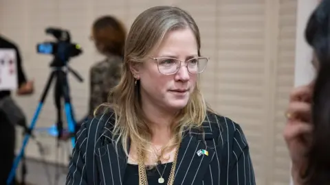 PA Media Dana Erlich has blond hair hair down past her shoulders and light-framed glasses. She is wearing a striped suit jacket with Israeli and Irish flag pins on it 