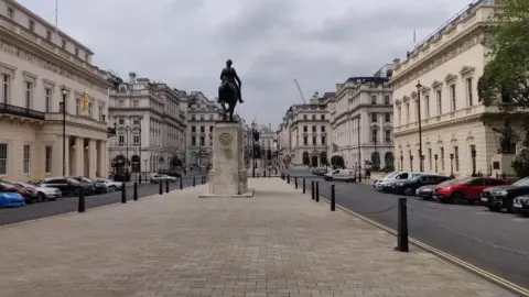 An equestrian statue on a stone plinth stands at the centre of Waterloo Place, with grand historic buildings lining both sides of the broad street in central London.