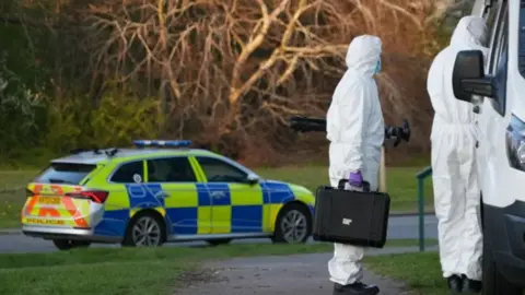 Two people in white hazmat outfits, one with a black briefcase, are standing next to a white van. There is a police car on the road to the left of the picture, with grass either side of the carriageway. There are trees in the background.
