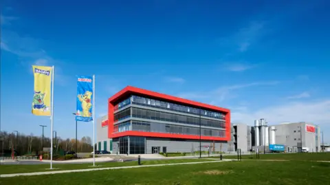 Haribo A modern industrial building with grey and red exterior panels displays a large “HARIBO” sign on the front. The site is surrounded by greenery, including grass, shrubs, and trees, with a road and gate leading up to the entrance.