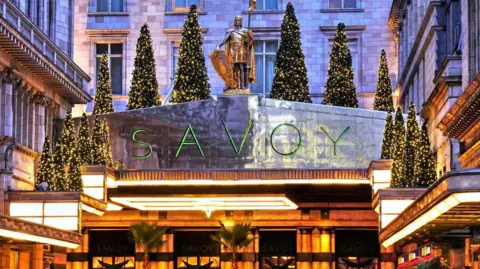 Getty Images Gold-coloured frontage of the Savoy hotel, with SAVOY in thin green letters above the entrance. There are fir trees above the SAVOY sign and a three-storey stone facade behind.