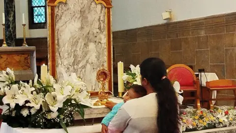 BBC Pilgrim holding a baby prays before the relic of Blessed Carlo Acutis