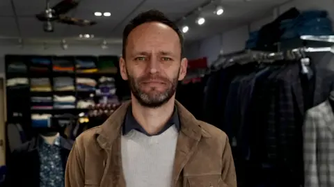 A headshot of Richard Dare. He is wearing a brown suede jacket, a cream jumper and a black shirt. He is looking at the camera and has a black beard and dark hair.