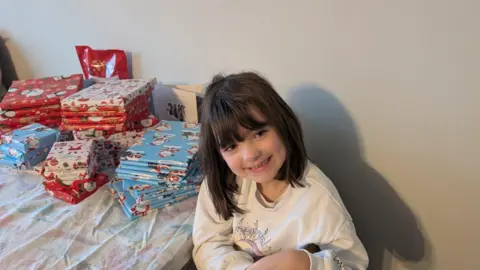 Kirsty Harper sat in front of the gifts on the table. She is smiling at the camera and has short black hair and is wearing a white top.