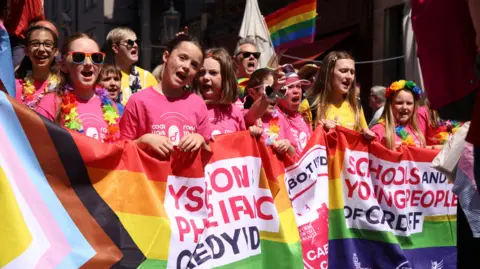 Jon Foster Cardiff Headshots A group of children wearing pink tops and colourful floral necklaces. They hold a LGBT+ flag. 