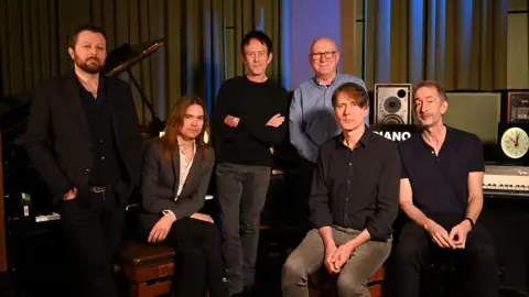 Mark Allan/BBC Six men, who form the band Suede, are pictured in front of amps, a piano and other musical equipment. They are dressed smartly with shirts and pants.