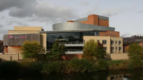 A large cream and glass-panelled theatre building with two grey balconies and a sign on the left hand side of the building saying: Theatre Severn. A river can be seen in the foreground and a tree-lined bank separates the river and theatre. 