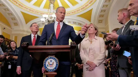 Getty Images Senate Majority Leader John Thune speaks to reporters at the US Capitol. He is accompanied by other senators and speaking behind a podium with the Senate logo.