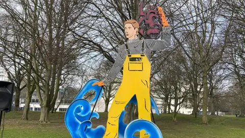 Jake Zuckerman / BBC The metal statue of Grim depicting a fisherman in yellow waterproof dungarees and a grey mariners sweater striding through blue waves carrying a red Havelock on his shoulder 