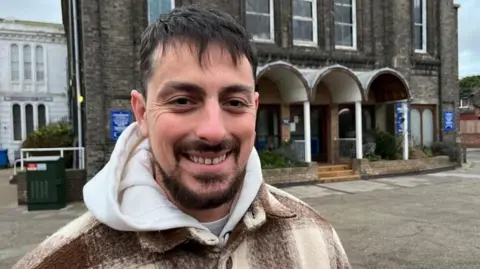Andrew Turner/BBC Ruben Cruz, who has a beard, moustache and dark hair, wearing a cream hooded top and brown and cream chequered jacket. He is standing outside the brick building. 