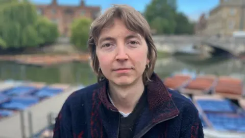 Phil Shepka/BBC A head-and-shoulders picture of William Ridgeon. He is standing alone in Cambridge city centre, with the river behind him. He has shoulder-length brown hair and is wearing gold earrings. He his wearing a blue zip-up top.