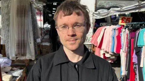 A man with light brown hair and glasses stands between rows of clothes.