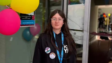 Poppy is a young woman with long, dark hair. She is wearing a black hoodie with two large badges on the front - one saying believe in yourself and the other a raincloud with a rainbow over the top. She is also wearing a blue lanyard and a pair of clear, framed glasses. She is stood in front of a large, glass window which has balloons attached to it.