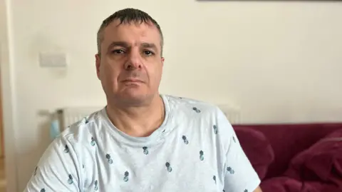 A middle-aged man looks at the camera. He is wearing a T-shirt and sitting on a sofa