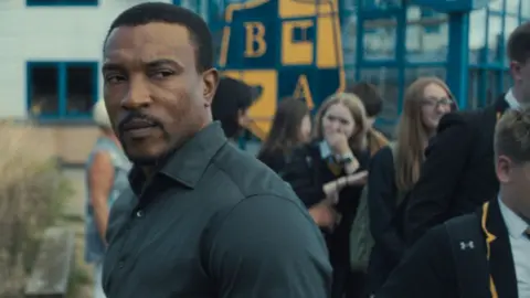 Netflix Ashley Walters with schoolchildren walking past him in a school yard in Adolescence