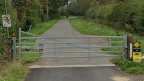 A large silver gate blocks the entrance to the bridleway, which is long and flanked by trees and bushes.