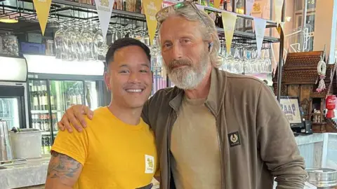 Mads Mikkelsen posing for a photo with a staff member at Zaap Thai. Mikkelsen is wearing a brown zip-up jumper. He has long grey hair. The staff member is wearing a yellow T-shirt. He has short dark hair and is smiling in the photo.