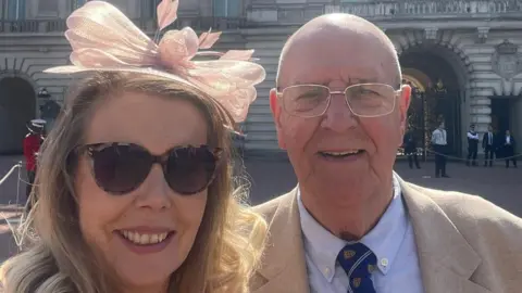 Roger Vine A male and female are taking a selfie - they are both smiling directly at the camera. The woman is blonde, wearing large tortoiseshell sunglasses and a pink fascinator. The male is bald, wearing thin silver glasses and a beige suit jacket with a blue shirt and navy tie.