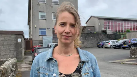 A woman with light brown hair blowing in the wind stands in front of a grey, functional school building with cars in the car park behind her and a grey sky above