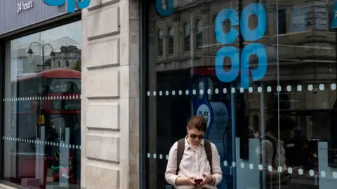 Reuters Man in shirtsleeves looking at phone walks in front of a Co-op store on The Strand, London