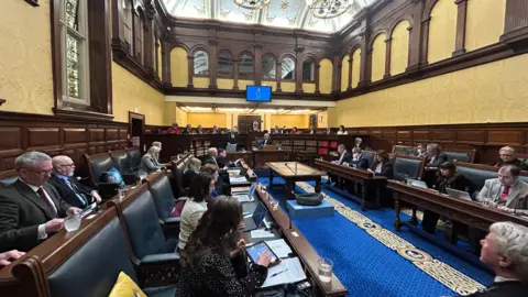 Members sitting in the Tynwald chamber, which has blue leather-clad wooden seat and desks and has blue carpet with gold decoration.