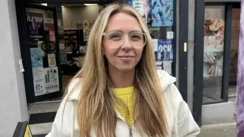 Leah Jones, who has long sandy blonde hair and clear-rimmed glasses, wears a white zip-up jacket and smiles at the camera, standing in front of a shop. It is a head and shoulders shot.