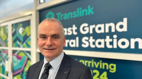 A man wearing a suit stands in front of a blue Translink sign