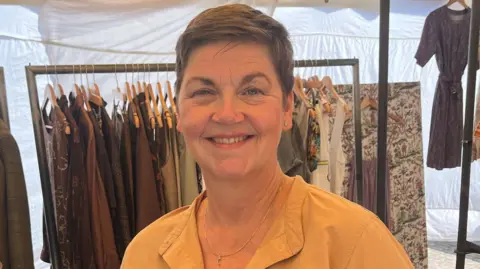 Ms Gregersen is smiling while standing at her stall. Behind her are metal clothing racks with clothes on wooden hangers. They are a range of designs in shades of brown, white cream and grey. She has short brown hair, blue eyes and is wearing a gold necklace with studded earrings. She is also wearing a yellow blouse. 