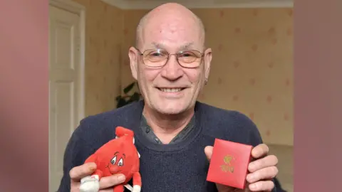 David Newton, wearing a grey sweater over a button-up T-shirt, and metal-framed spectacles, smiles as he holds up a red presentation box embossed with a gold National Blood Service logo and the figure "100" in one hand. In his left ear is an earring. In the other hand is a red soft toy mascot of a blood drop. He is inside what looks like a living room. 