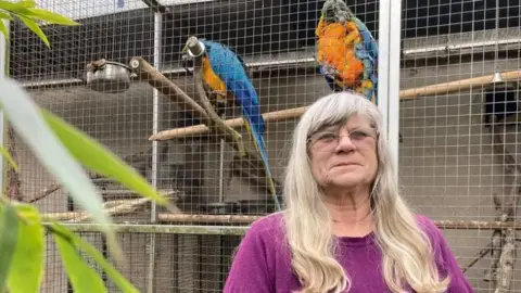 A woman in her mid 70s with grey hair that extends past her shoulders. She is wearing a purple top. Behind her are two parrots in cages.