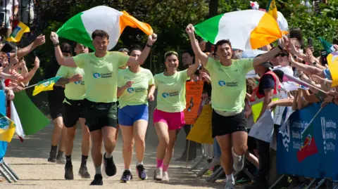 PA Media Jordan and Cian run towards the finish line of their Ireland challenge wearing florescent green tops and black running shorts. They are both carrying an Irish flag each while a crowd of runners in matching outfits run behind them. On either side of them is a crowd of people cheering them on.
