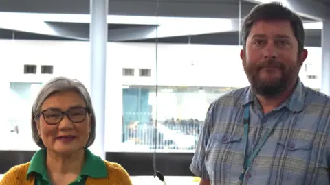 Suffolk New College Kin Alderson wearing a mustard coloured cardigan over a green polo shirt and glasses. She is smiling. Standing next to her is Kristian Sanderson, who is wearing a short-sleeved shirt and a light blue lanyard. He is looking into the camera. 