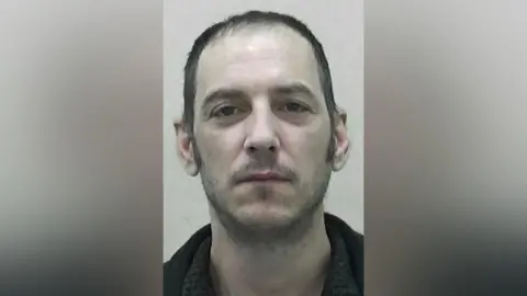 Mugshot of Mark Harvey. He has pale, pasty skin, scruffy short dark hair and a faint wispy dark beard