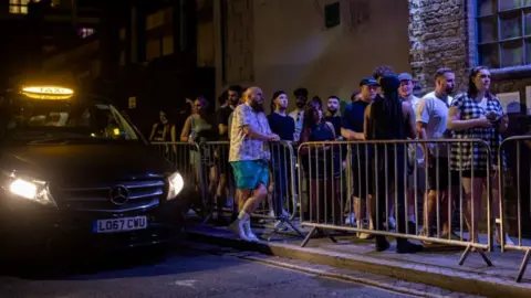 Getty Images A stock image showing a black taxi pulling up alongside metal barriers outside a bar, where lots of people are queuing to get inside.
