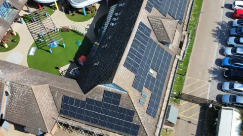 An aerial drone shot of the hospice roof and three sections covered in black solarpanels. There is a car park to the right and a courtyard in the middle with green areas and a pergola.