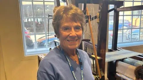 BBC A woman with short brown hair is pictured wearing a blue knitted jumper with a blue lanyard with wording that reads "volunteer" in white capital letters. She is stood in front of a window and also next to a wooden weaving loom. 