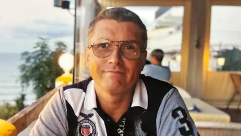 A man with short dark grey hair and wearing glasses and a black and white polo top is smiling at the camera. He is sitting in a restaurant with a sea view behind him.