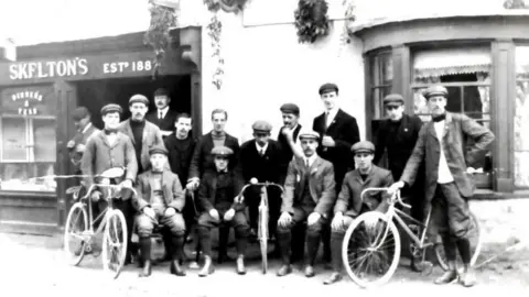 An old black and white photo of men in flat caps posing with bicycles. 