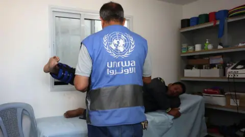 Getty Images An Unrwa worker successful Khan Younis, Gaza. He wears a bluish Unrwa vest and is treating a man wearing a limb brace. 