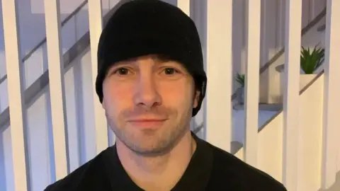 Shelley Martin Barry Martin is smiling at the camera. He is wearing a black top and a black beanie hat and is standing in front of stairs in a house